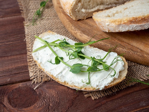 Nahaufnahme eines Stilllebens aus frischem knusprigem Brot Eine Scheibe Brot, die mit Hüttenkäse bestrichen und mit Mikrogrün verziert ist Leinenserviette braun Holzhintergrund Draufsicht flach gelegt