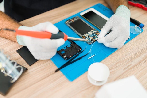 Nahaufnahme eines Technikers mit Handschuhen, der das Motherboard eines alten Smartphones an seinem Werkstatttisch lötet und repariert