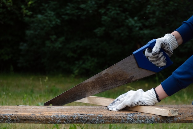 Nahaufnahme eines Tischlers mit Handschuhen und Handsägen von Holzbrettern mit alter stumpfer Säge im Garten.