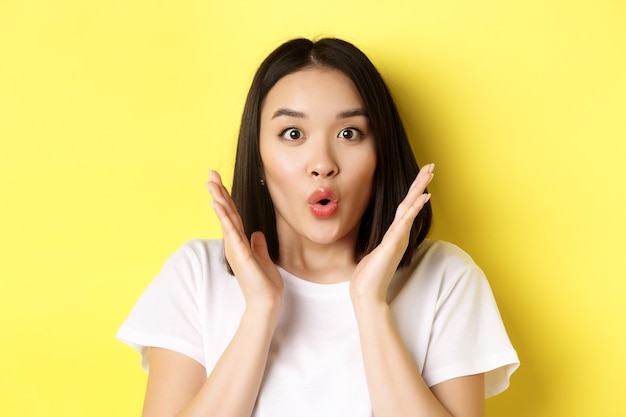 Nahaufnahme eines überraschten asiatischen Mädchens sagen wow, starren in die Kamera amzed mit den Händen in der Nähe des Gesichts und stehen auf gelbem Hintergrund.
