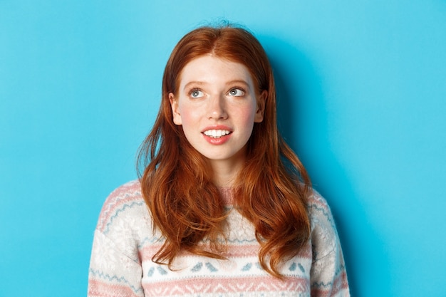 Nahaufnahme eines verträumten Teenie-Mädchens mit roten Haaren, das die obere linke Ecke betrachtet und lächelt, vor blauem Hintergrund stehend.