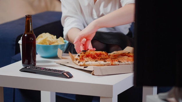 Nahaufnahme eines weißen Tisches mit ungesundem Essen und Bier im Wohnzimmer. Junge Frau isst ein Stück Pizza aus der Lieferbox, die auf der Couch im Fernsehen sitzt. Person mit Essen zum Mitnehmen zu Hause