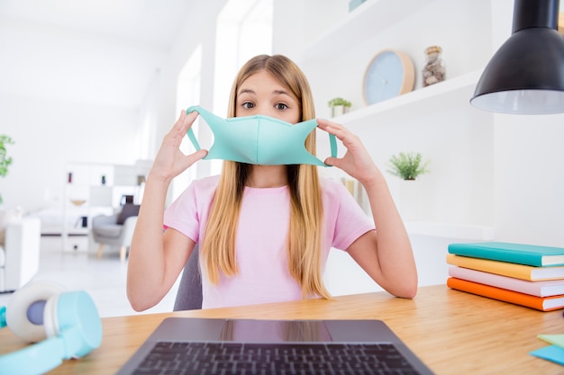 Nahaufnahme Foto von schockiertem Schüler Kind kleines Mädchen sitzen Schreibtisch Studie Remote-Laptop haben Online-Kommunikation Gespräch Lehrer sprechen sprechen medizinische Maske im Haus drinnen tragen