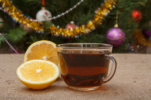 Nahaufnahme Glastasse Tee und Zitronenstücke auf dem Tisch mit Sackleinen und Weihnachtsbaum mit Spielzeugbällen und Girlanden im Hintergrund.