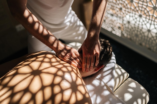 Nahaufnahme hochwinkelansicht von unerkennbarem Masseur männlich, der therapeutische Halsmassage an einem Mann macht, der auf einer Massagesofa auf dem Hintergrund von Sonnenlicht und wunderschönen Schatten liegt Konzept der Ruhe mit Spa-Behandlung