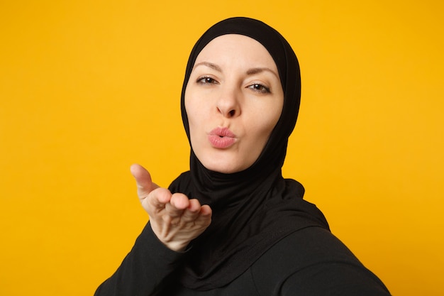 Nahaufnahme junge arabische muslimische frau in hijab schwarzer kleidung, die selfie auf dem handy isoliert auf gelbem wandporträt macht. Menschen religiöses Lifestyle-Konzept.