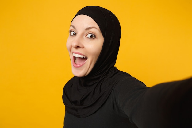 Nahaufnahme junge arabische muslimische frau in hijab schwarzer kleidung, die selfie auf dem handy isoliert auf gelbem wandporträt macht. Menschen religiöses Lifestyle-Konzept.