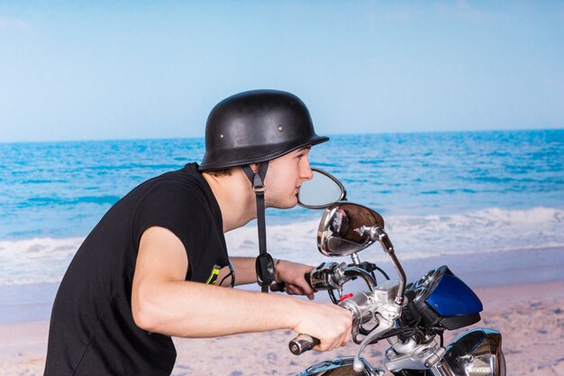 Nahaufnahme junger gutaussehender Mann, der ein Motorrad am Strand mit schwarzem Helm auf einem tropischen Klima reitet.