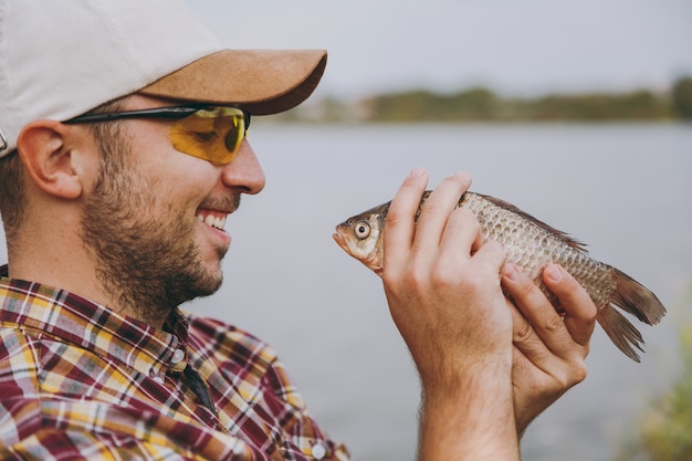 Nahaufnahme Junger unrasierter lächelnder Mann in kariertem Hemd, Mütze und Sonnenbrille hat einen Fisch gefangen und sieht ihn am Ufer des Sees auf dem Hintergrund des Wassers an. Lifestyle, Erholung, Freizeitkonzept für Fischer