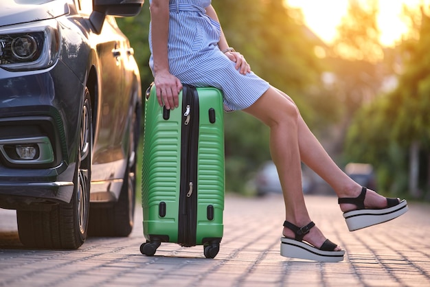 Nahaufnahme junger weiblicher Beine, die auf einer Koffertasche neben dem Auto sitzen Reise- und Urlaubskonzept