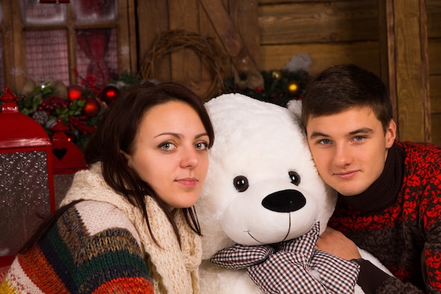 Nahaufnahme junges weißes Paar in Winter-Outfits umarmt weiße Bärenpuppe beim Blick in die Kamera.