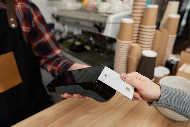 Foto nahaufnahme, kundenhand, die kreditkarte in der nähe des nfc-terminals hält. barista akzeptiert zahlungen per kreditkarte über den pos-automaten im café