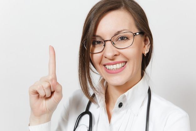 Nahaufnahme lächelnde Arzt Frau mit Stethoskop, Brille isoliert auf weißem Hintergrund. Ärztin in medizinischer Uniform, die Zeigefinger auf Kopienraum zeigt. Gesundheitspersonal, Medizinkonzept.
