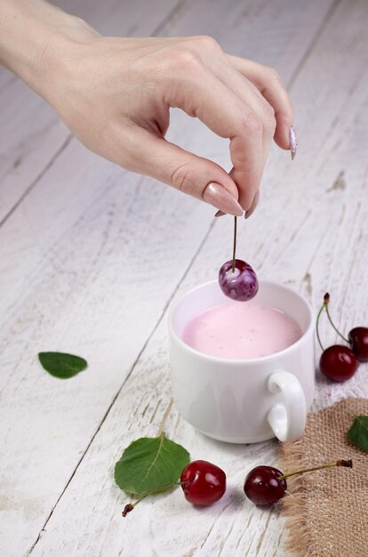 Nahaufnahme Mädchenhände halten frische Kirsche über Glas mit Joghurt, heller Holztisch. Leckeres Frühstück und ein guter Start in den Tag