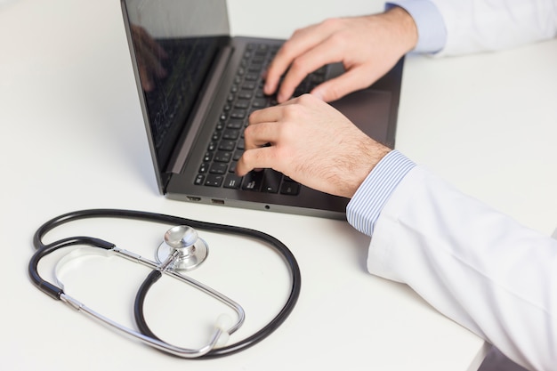 Nahaufnahme männlichen Doktors, der Laptop über weißem Schreibtisch mit Stethoskop verwendet