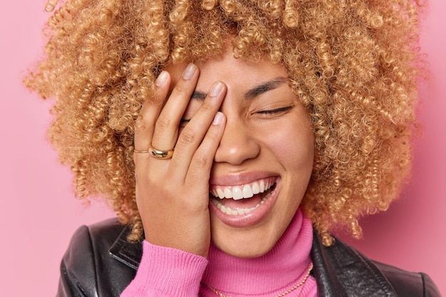 Nahaufnahme Porträt einer glücklichen aufrichtigen Frau bedeckt die Hälfte des Gesichts mit der Handfläche hält die Augen geschlossen Lächeln drückt im Großen und Ganzen positive Emotionen aus Lachen über etwas Posen im Innenbereich vor rosafarbenem Studiohintergrund