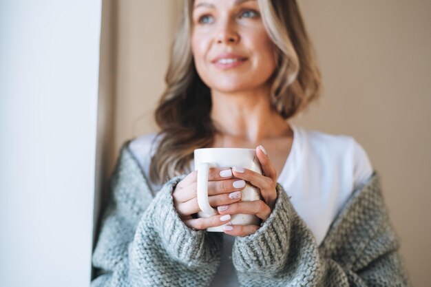 Nahaufnahme Porträt einer jungen schönen Frau vierzig Jahre mit blonden langen lockigen Haaren in einem gemütlichen grauen Strickpullover mit einer Tasse Tee in den Händen im hellen Interieur zu Hause