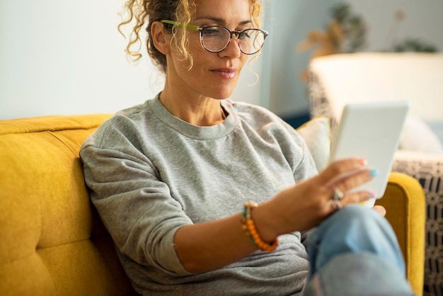 Nahaufnahme Porträt einer modernen Frau, weibliche Erwachsene, die ein Buch von einem elektronischen Lesegerät lesen. Dame mit Brille und Lächeln, die entspannte Zeit zu Hause auf dem Sofa genießen