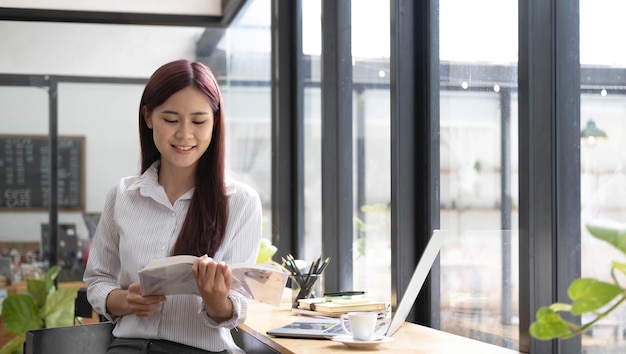 Nahaufnahme Porträt einer schönen jungen asiatischen Frau, die lächelt und ein Buch im Café betrachtet