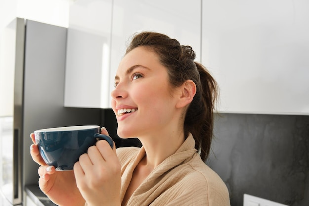 Nahaufnahme Porträt einer verträumt lächelnden jungen Frau, die Kaffee trinkt und nachdenklich aussieht, steht in der Küche