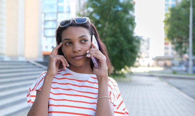 Nahaufnahme Porträt eines attraktiven, stilvollen, dunkelhäutigen Mädchens mit Afro-Frisur, das auf dem Handy isoliert auf Straßenhintergrund spricht