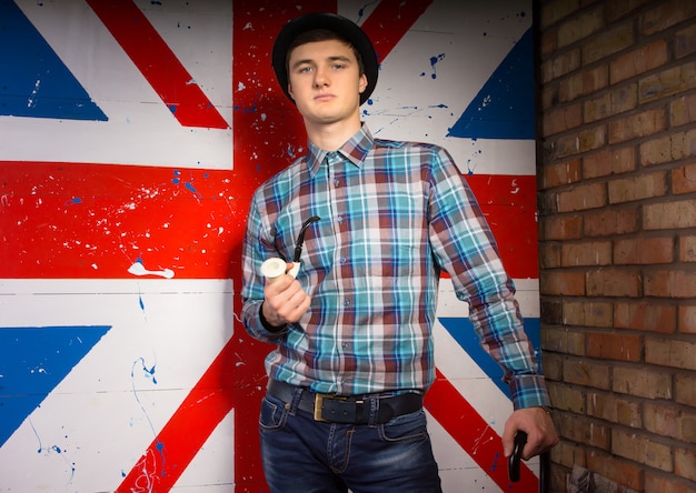 Nahaufnahme Porträt eines wunderschönen jungen Mannes in trendigem Outfit mit Tabakpfeife und Stock vor dem britischen Flaggendruck beim Blick in die Kamera.