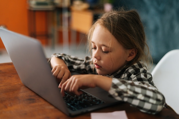 Nahaufnahme Seitenansicht eines hübschen elementaren Kindermädchens, das das Tippen auf der Tastatur des Laptops verwendet, der am Schreibtisch sitzt