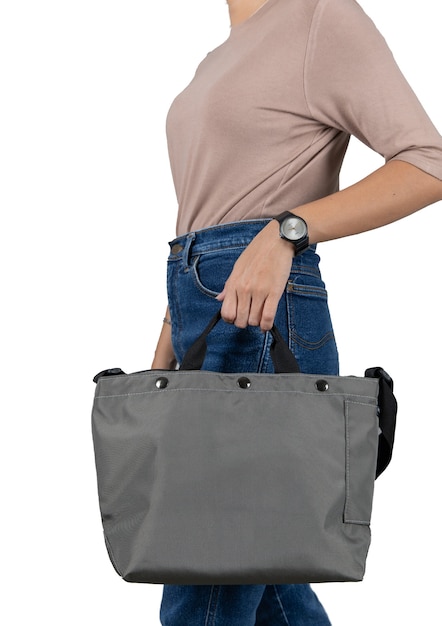 Nahaufnahme Studioaufnahme des weiblichen Modells in braunem Langarmhemd und Jeans stehen mit Griff der modischen trendigen urbanen großen blauen Unisex-Handtasche mit langem Schultergurt vor grauem Hintergrund.