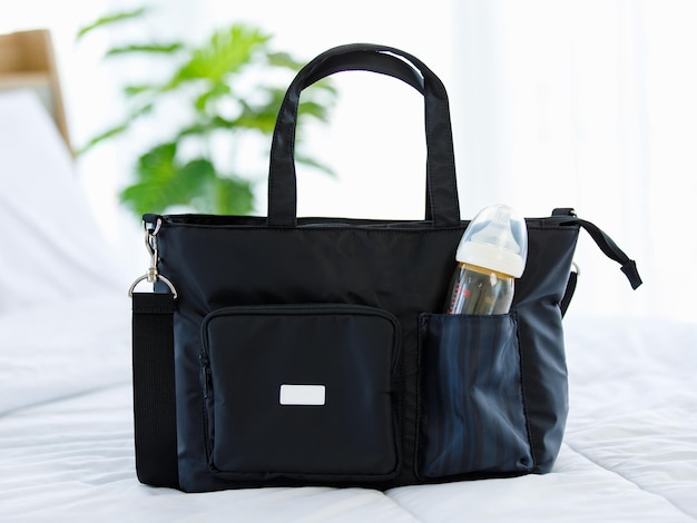 Nahaufnahme-Studioaufnahme einer schwarzen Multifunktions-Mehrzweck-Utility-Zubehör-Handtasche für Neugeborene mit Milch-Babyflasche und Plastikkappe in einer elastischen Tasche mit Griff und Riemen auf dem Bett
