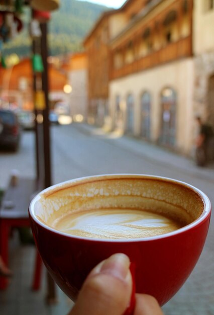 Nahaufnahme Textur einer halben Tasse heißen Cappuccino-Kaffees in der Hand der Frau gegen verschwommenen Altstadt