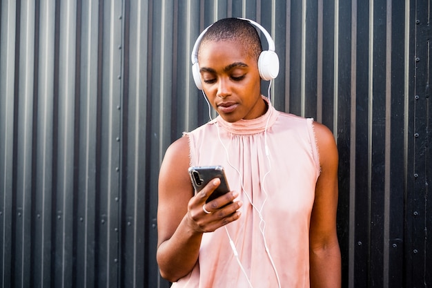 Nahaufnahme und Porträt einer schwarzen und afroschönen Frau, die ihr Telefon benutzt, während sie mit weißen Kopfhörern Musik hört - schwarze Wand im Hintergrund - die Technologie liebend