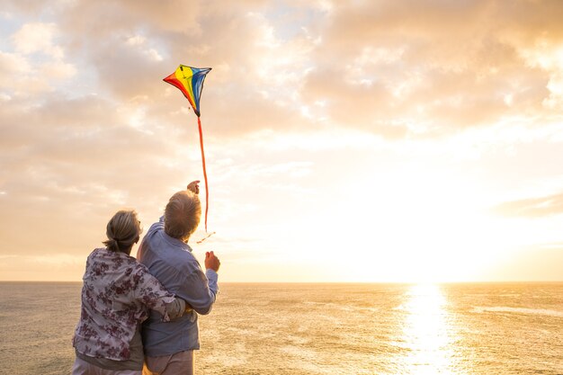 Nahaufnahme und Porträt von zwei alten und reifen Menschen, die mit einem Drachen am Strand mit dem Meer im Hintergrund mit Sonnenuntergang spielen und genießen - aktive Senioren, die Spaß haben