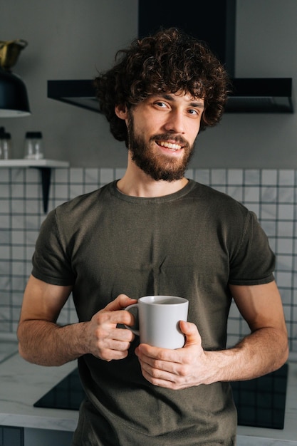 Nahaufnahme vertikales Porträt eines lächelnden jungen Mannes, der in der Hand eine Tasse mit Morgenkaffee hält und in die Kamera blickt, die an der Küchentheke steht