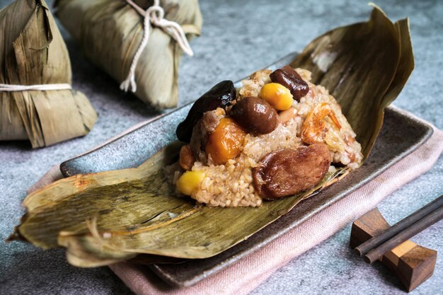 Nahaufnahme von asiatischen leckeren hausgemachten Lebensmitteln im Drachenboot (duan wu) Festival, Reisknödel oder Zongzi, eingewickelt durch getrocknete Bambusblätter auf Teller mit Tee auf schwarzer Oberfläche