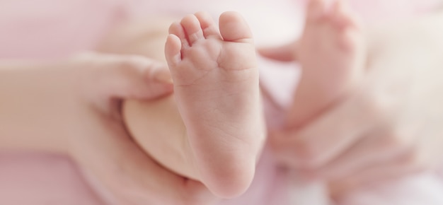 Nahaufnahme von den Mutterhänden, die nette kleine Babyfüße halten und Babyfuß zeigen. Rosa hintergrund. Horizontal.