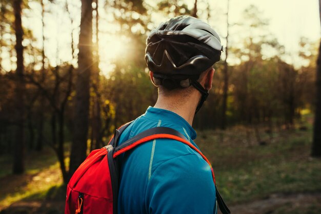 Nahaufnahme von der Rückseite eines gutaussehenden jungen bärtigen Radfahrers mit Schutzhelm, blauem T-Shirt und rotem Rucksack, der wegschaut und über Route und Radfahren nachdenkt Reisekonzept Sport-Lifestyle