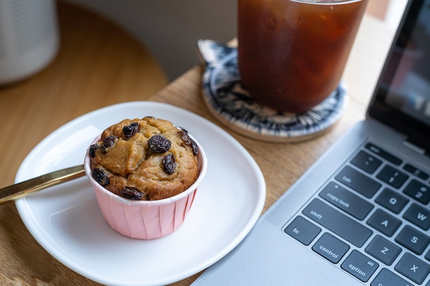 Nahaufnahme von Eiskaffee in Tassenbecher und hausgemachtem Bananen-Tassenkuchen mit Tastatur-Laptop-Computer auf dem Schreibtisch aus Holz im Café im Café, während des Geschäftskonzepts