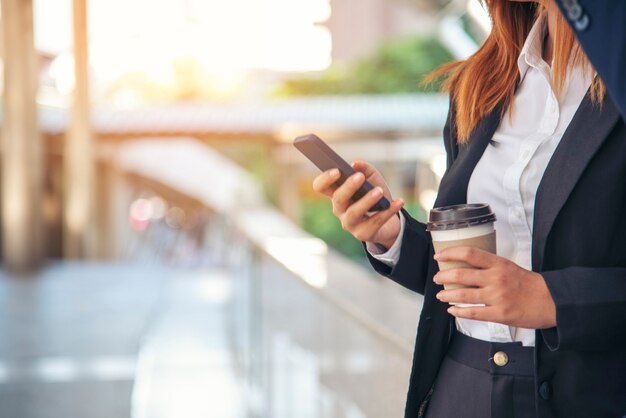 Nahaufnahme von Frauenhänden, die Kaffeetasse in der Stadt mit Smartphone außerhalb des Büros in der modernen Stadt halten