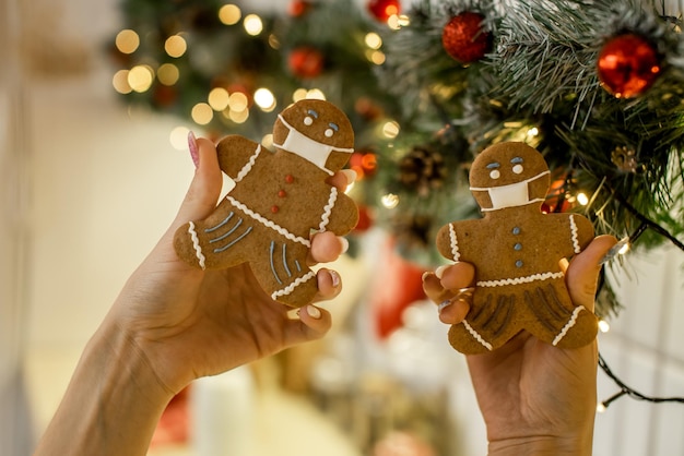 Nahaufnahme von Frauenhänden, die Lebkuchenmann und -frau in Gesichtsmaske auf Weihnachtsbaumhintergrund halten