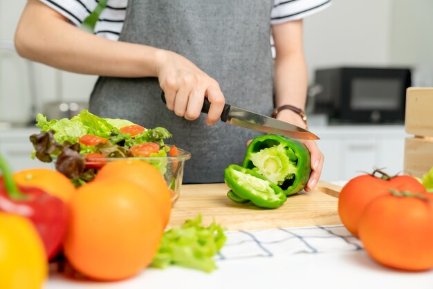 Nahaufnahme von Frauenhänden verwenden ein Messer, um die Paprika und verschiedene grüne Blattgemüse zu schneiden