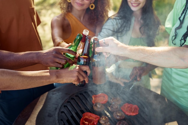 Nahaufnahme von Freunden, die Bierflaschen über dem Grill klirren, während sie im Sommer eine Grillparty im Freien genießen