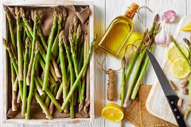 Nahaufnahme von frischem grünem Spargel in einer rustikalen Holzkiste und auf einem Schneidebrett mit Messer, Zitronenscheiben und Flasche Olivenöl