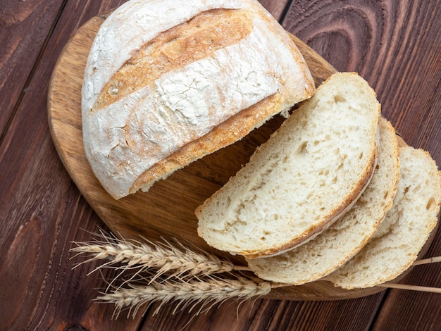 Nahaufnahme von frischem knusprigem Weizenbrot in Scheiben geschnitten liegt auf einem Holzbrett Hölzerner brauner Hintergrund Mehrere Ähren liegen neben dem Brot Draufsicht flach gelegt