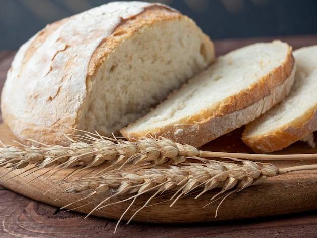 Nahaufnahme von frischem knusprigem Weizenbrot in Scheiben geschnitten liegt auf einem Holzbrett Holzbrauner Hintergrund Mehrere Ähren liegen neben dem Brot Seitenansicht