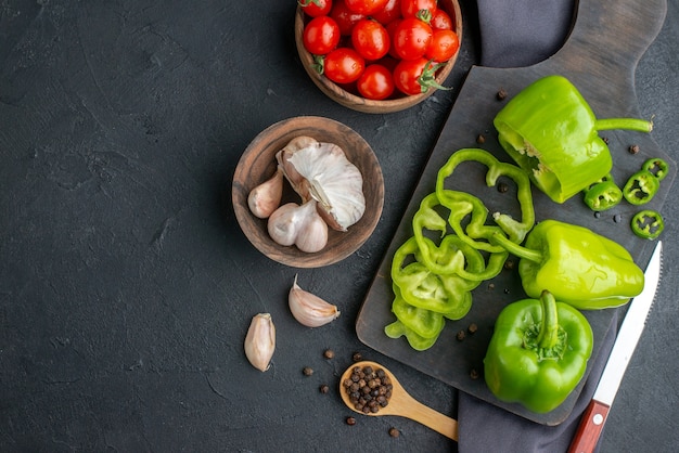 Nahaufnahme von ganzen geschnittenen gehackten grünen Paprika auf Holzbrett Tomaten in Schüssel Knoblauch auf dunklem Handtuch auf schwarzer Oberfläche
