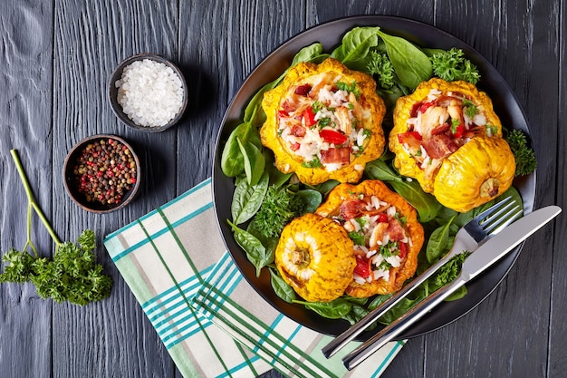 Nahaufnahme von gebackenem Pattypan-Kürbis gefüllt mit Reis, gebratenem Hühnerfleisch, knusprig gebratenem Speck, rotem Paprika und serviert mit Spinatblättern und Petersilie auf einem schwarzen Teller mit Gabel und Messer