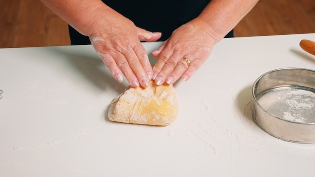 Nahaufnahme von Händen der älteren Bäckerei, die Teig knetet. Pensionierter älterer Bäcker mit Knochen, der Zutaten mit gesiebtem Weizenmehl mischt, das sich auf einer bemehlten Oberfläche zum Backen von traditionellem Kuchen, Brot bildet