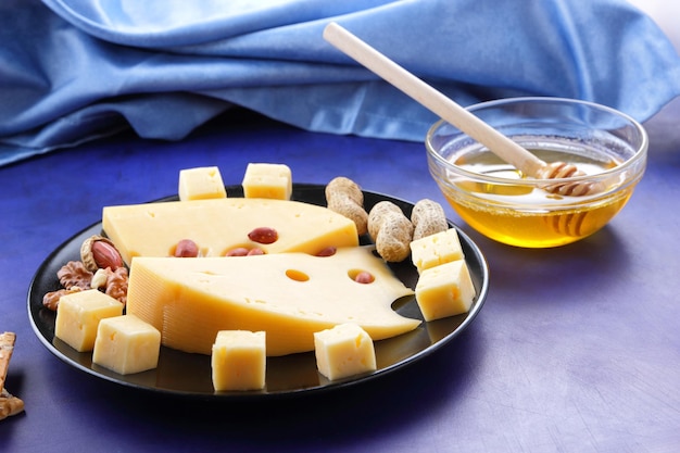 Nahaufnahme von Hartkäsescheiben mit großen Löchern und einer blauen Serviette auf blauem Hintergrund Scheiben harter gelber Käse mit Nüssen, Honig und Brötchen auf einem schwarzen Teller