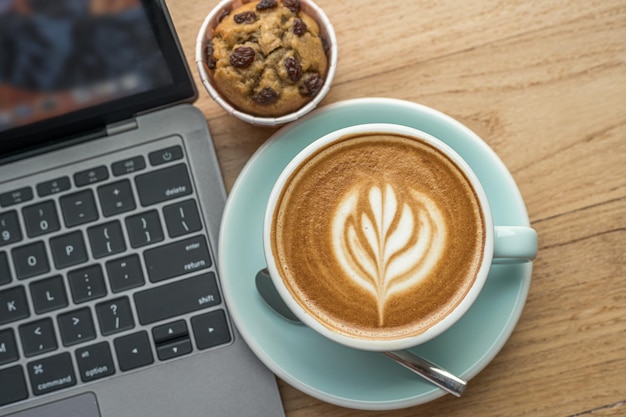 Nahaufnahme von heißem Kaffee Latte mit Latte-Art-Milchschaum im Becher und hausgemachtem Bananencup-Kuchen mit Laptop-Computer auf Holzschreibtisch-Schreibtisch im Café im Cafeduring-Geschäftsarbeitskonzept