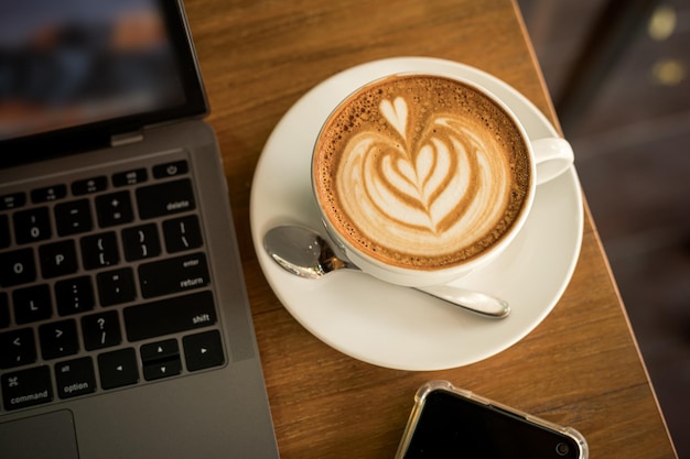 Nahaufnahme von heißem Kaffee Latte mit Latte-Art-Milchschaum in Tassenbecher und Laptop-Computer und Smartphone auf Holzschreibtisch Schreibtisch im Café im Cafeduring-Geschäftsarbeitskonzept